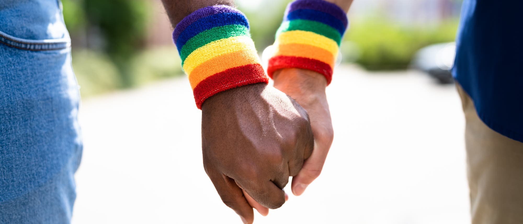 Ein schwules Paar hält einander an den Händen, am Handgelenk tragen sie Regenbogen-Bänder