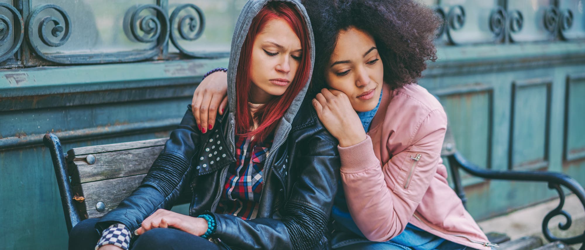 Zwei junge Frauen spenden einander Trost. Die Frau links trägt eine schwarze Lederjacke und hat rot getönte Haare, die Frau rechts trägt einen rosa gefärbten Pullover, blaue Jeans und hat dunkle Locken. Beide sitzen auf einer Bank.