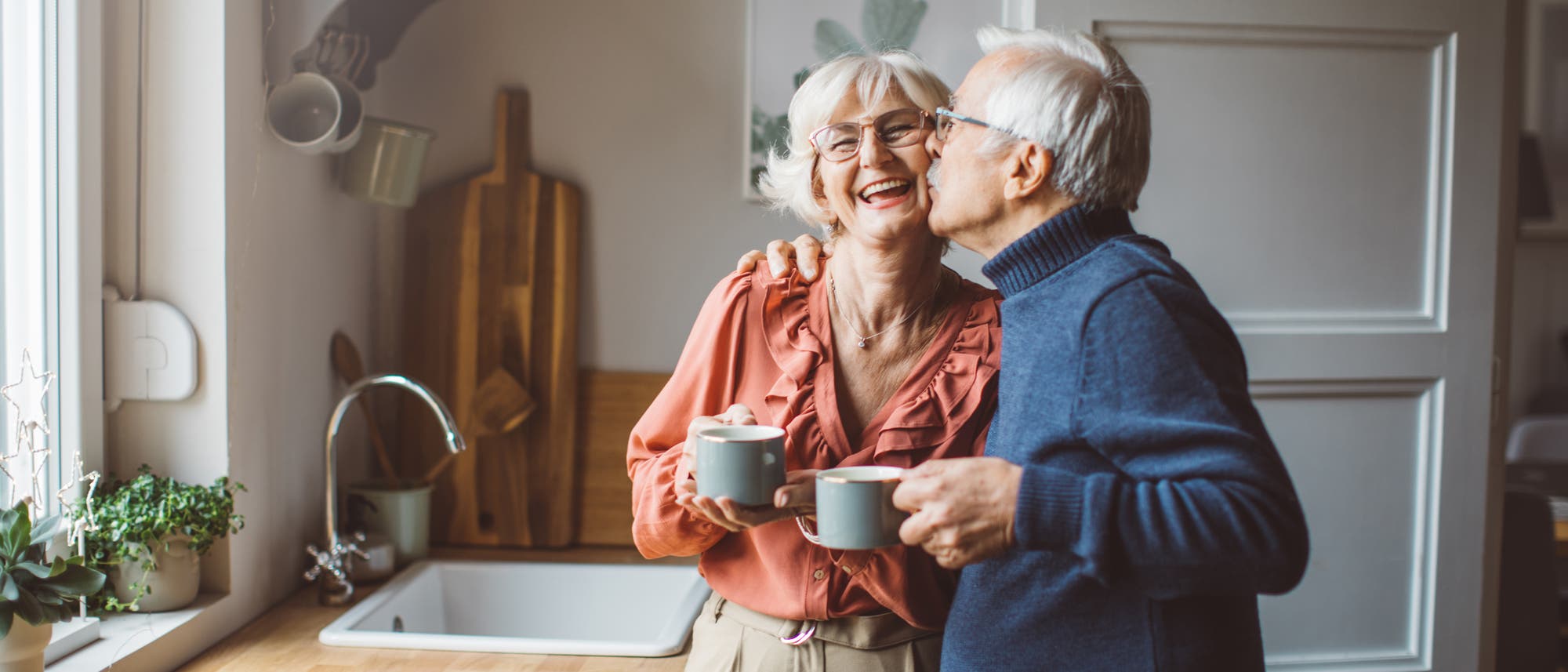 Ein älteres Paar steht zusammen in der Küche mit Tassen in der Hand und der Mann gibt der Frau einen Kuss auf die Wange.