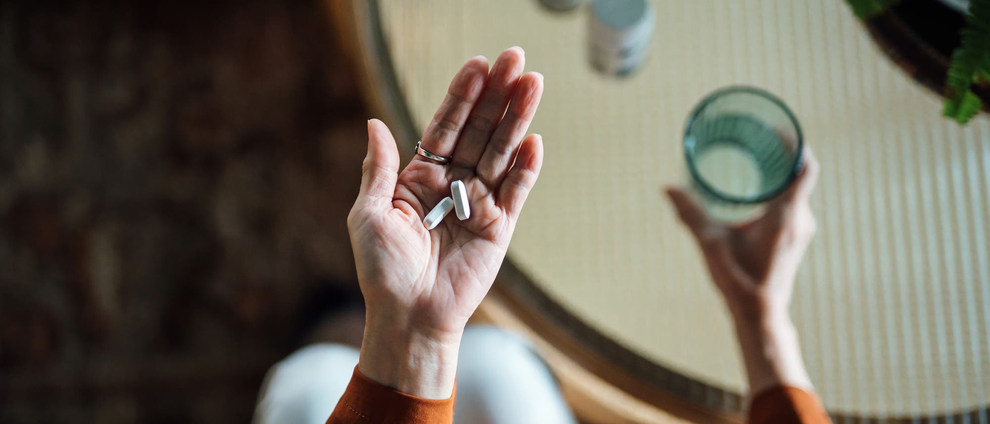Überkopfansicht einer Frau, die Medikamente in der Hand hält. Auf dem Tisch steht ein Glas Wasser. 