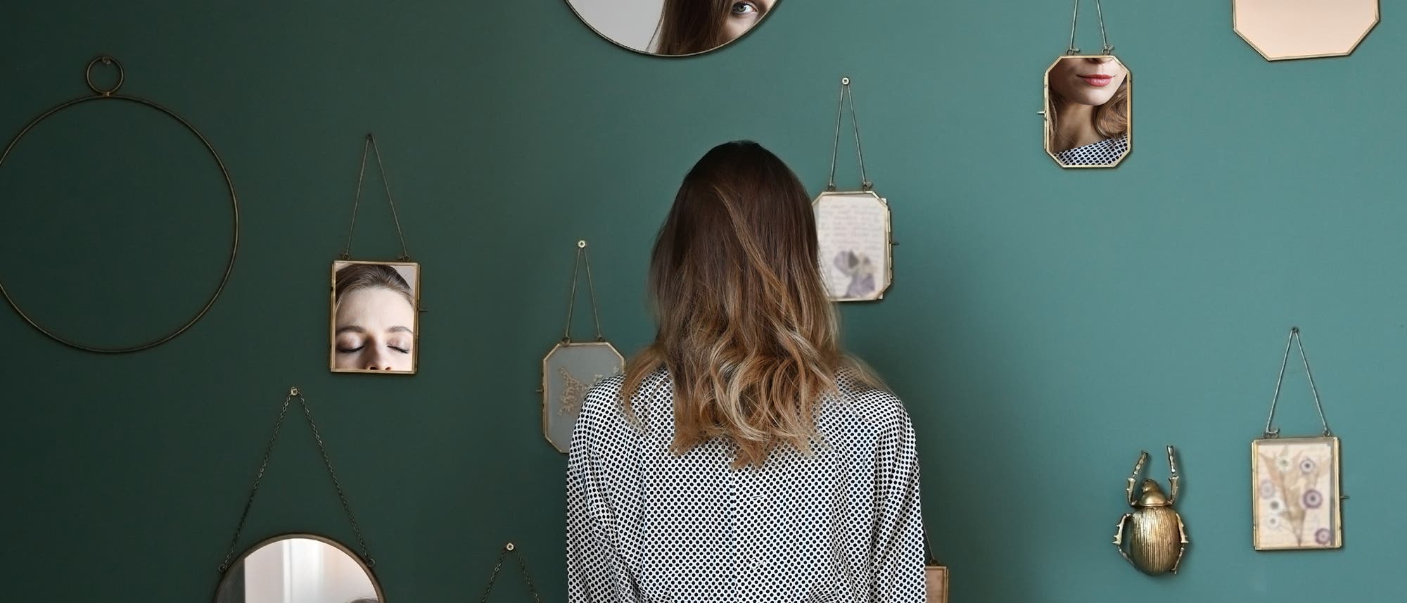 Frau von hinten, an der Wand viele verschiedene Spiegel, in denen Teile ihres Gesichts zu sehen sind