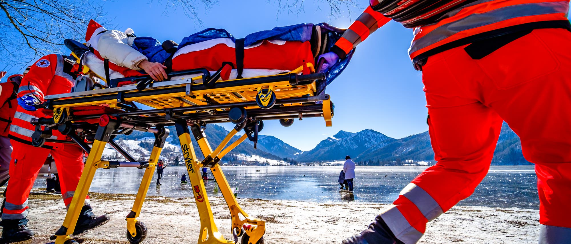 Zwei Sanitäter transportieren eine Person auf einer Trage an einem zugefrorenen See.