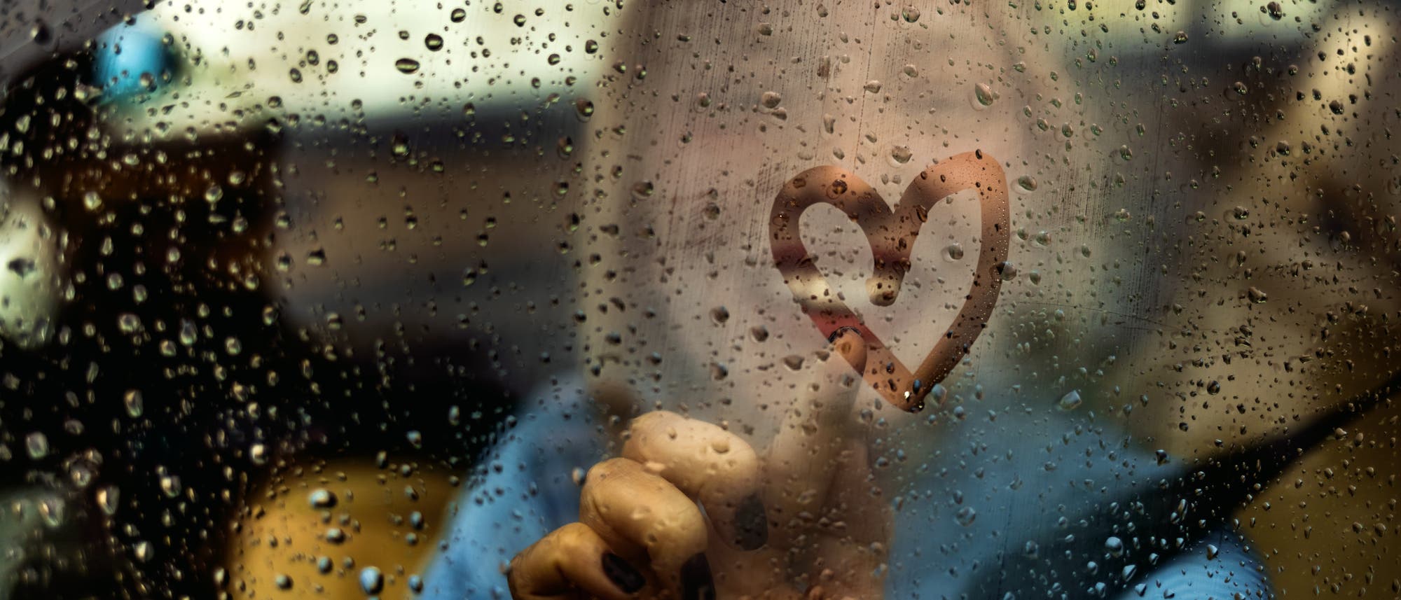 Herz, das mit dem Finger auf eine verregnete Autoscheibe gemalt wurde. Im Hintergrund ist verschwommen die Frau zu erkennen, die das Herz gemalt hat. 