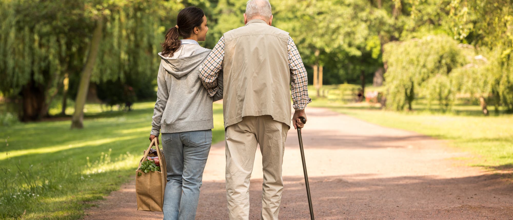 Eine junge Frau und ein &auml;lterer Mann gehen gemeinsam auf einem Parkweg spazieren. Die Frau tr&auml;gt eine Einkaufstasche und h&auml;lt den Arm des Mannes, der einen Gehstock benutzt. Umgeben von gr&uuml;nen B&auml;umen und einem sonnigen Himmel, vermittelt die Szene ein Gef&uuml;hl von Unterst&uuml;tzung und Gemeinschaft.