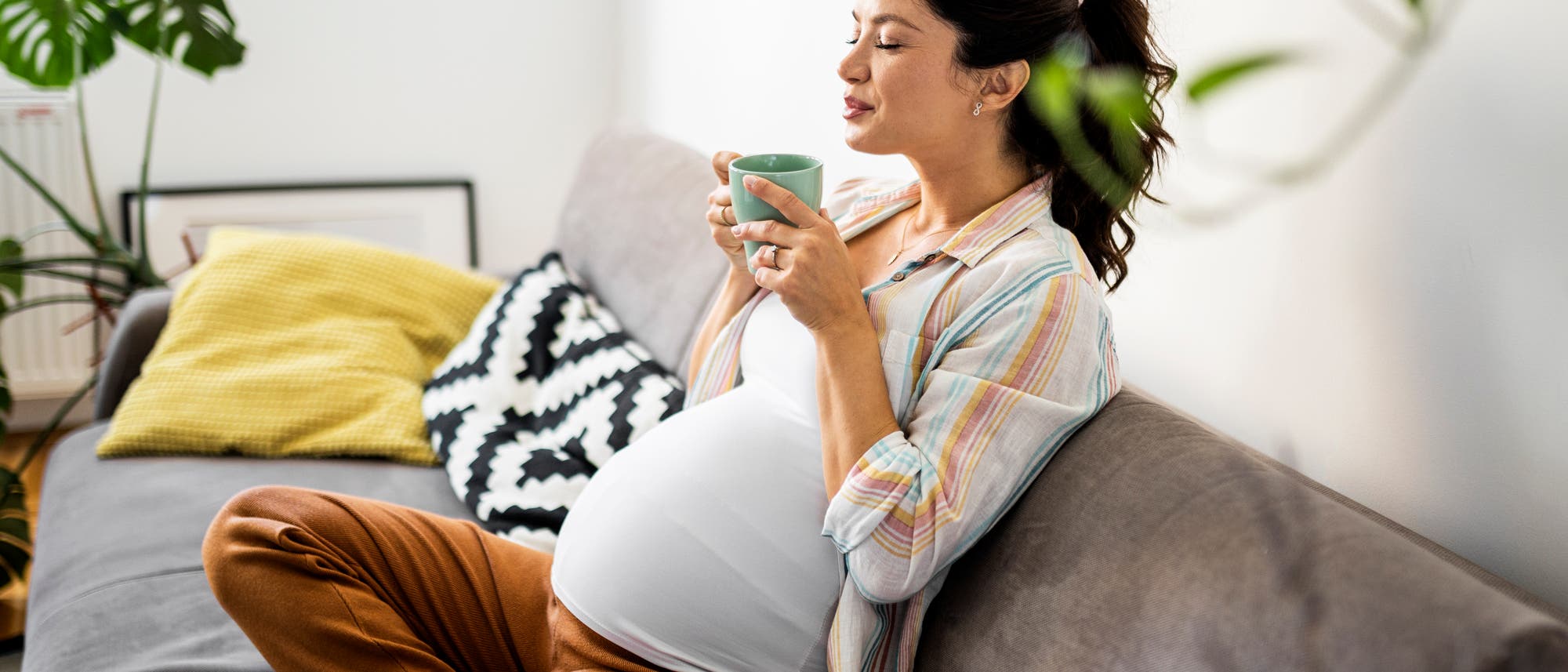 Schwangere sitzt auf Sofs und riecht an Tasse