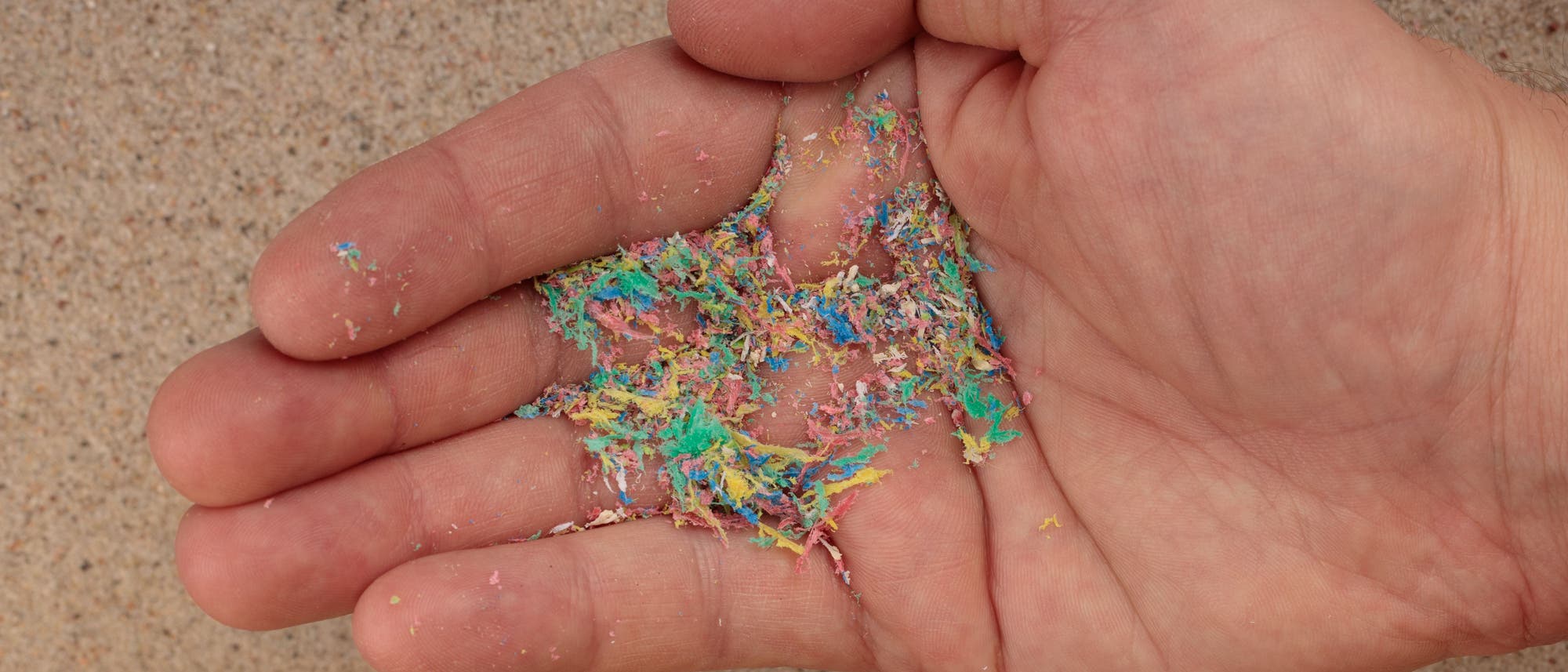 Eine Hand vor einem Strand, in der Hand liegen bunte Fasern aus Mikroplastik