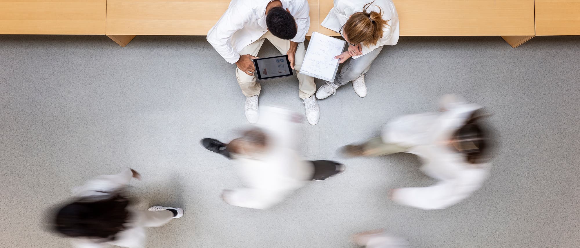 Zwei sitzende Medizinstudenten besprechen sich, andere laufen an ihnen vorbei, Blick von oben aus der Vogelperspektive