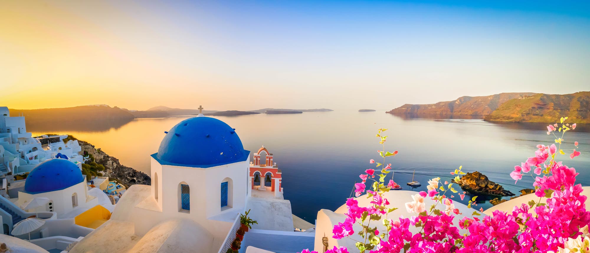 Santorini gilt als Inbegriff griechischer Idylle: Weeiße Häuser strahlen im warmen Abendlicht, der Blick geht auf die Bucht mit tiefblauem Wasser. Im Vordergrund sind rote Blumen