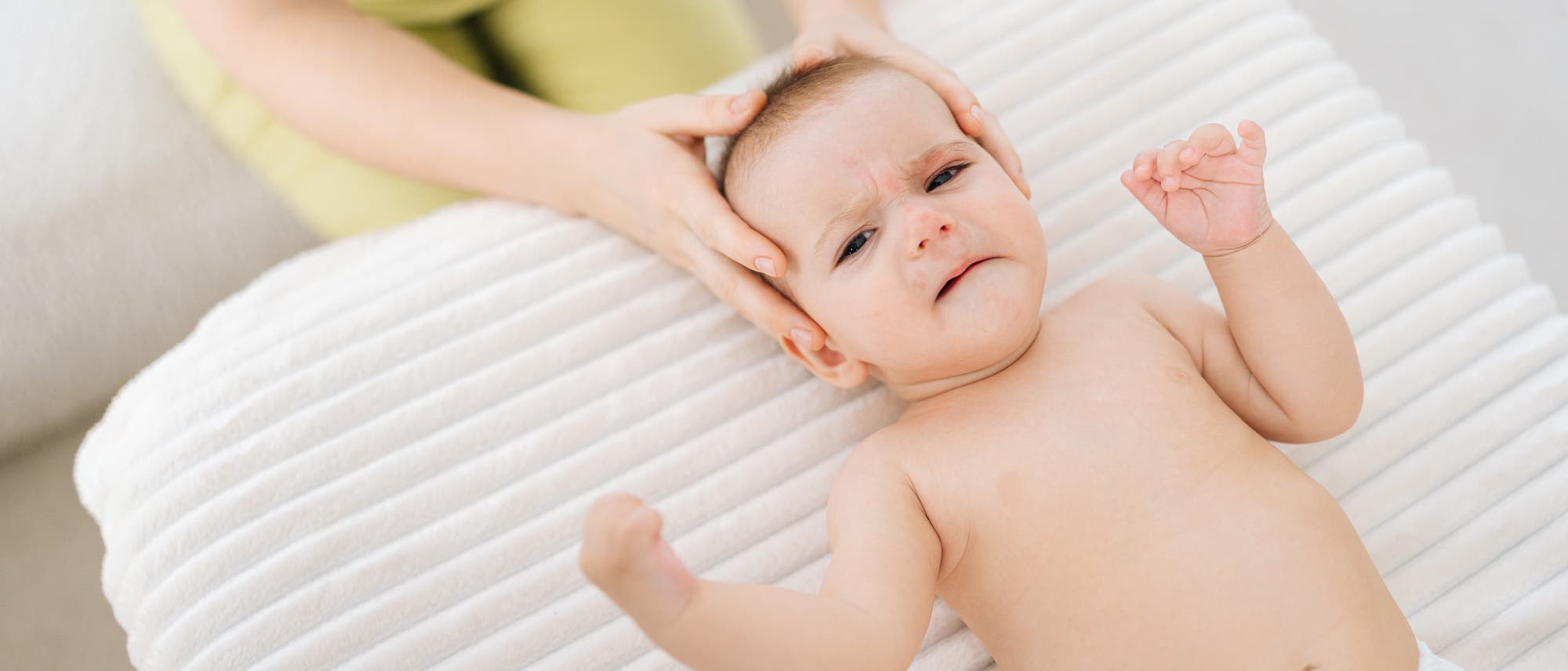 Ein Baby liegt auf einer Behandlunsgliege und wird von einem Osteopathen mit beiden Händen am Kopf angefasst 