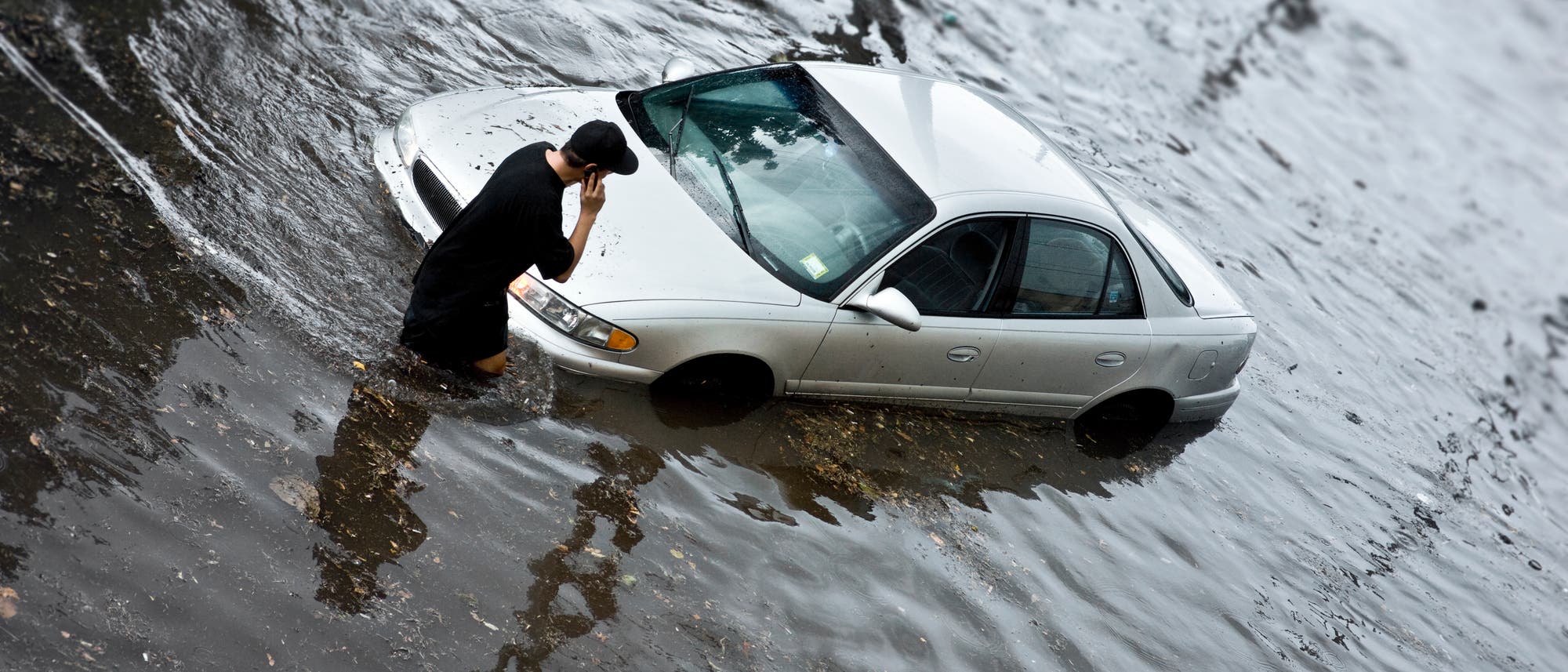 Mann steht mit Handy am Ohr knietief im Wasser vor seinem Auto nach der Flutkatastrophe.