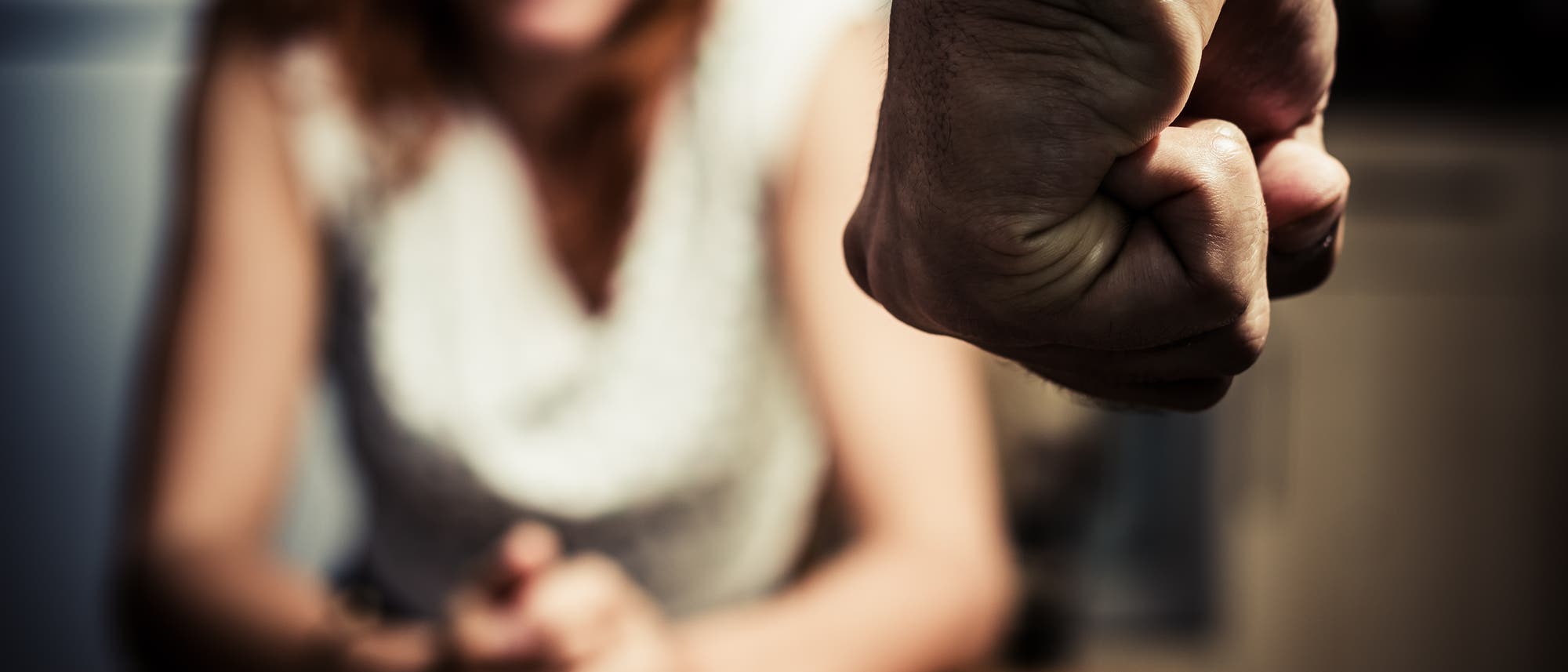 Eine unscharfe Person sitzt mit gefalteten Händen am Tisch, während im Vordergrund eine geballte Faust sichtbar ist. Die Szene vermittelt eine angespannte und bedrohliche Atmosphäre.