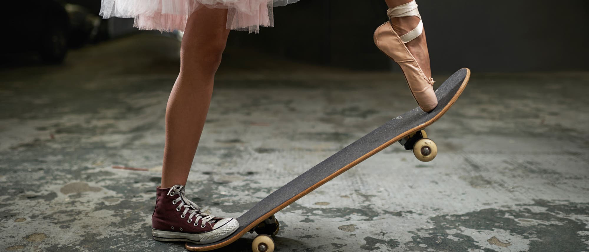 Junge Frau auf einem Skateboard, die am rechten Fuß einen Turnschuh und am linken Fuß einen Ballettschuh trägt.