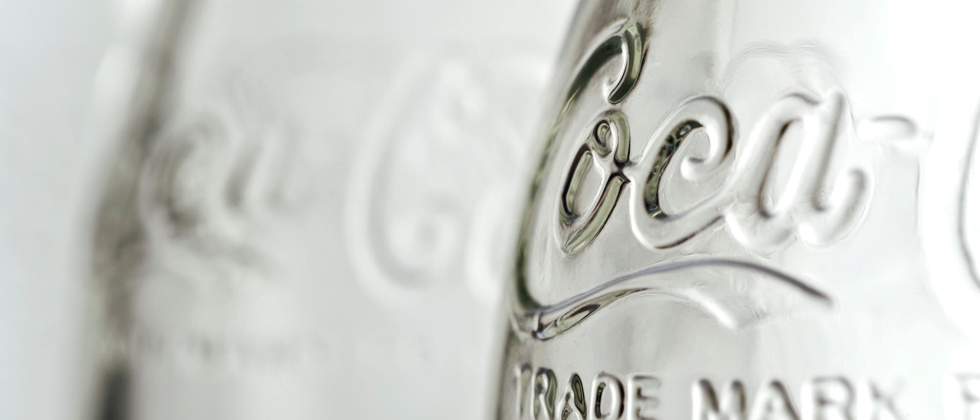 Eine Nahaufnahme von zwei leeren Glasflaschen mit dem eingravierten Schriftzug "Coca-Cola" und "TRADE MARK". Die Flaschen sind transparent und reflektieren das Licht, wodurch der Schriftzug hervorgehoben wird. Der Fokus liegt auf der vorderen Flasche, während die hintere Flasche unscharf ist. Eine Nahaufnahme von zwei leeren Glasflaschen mit dem eingravierten Schriftzug "Coca-Cola" und "TRADE MARK". Die Flaschen sind transparent und reflektieren das Licht, wodurch der Schriftzug hervorgehoben wird. Der Fokus liegt auf der vorderen Flasche, während die hintere Flasche unscharf ist.