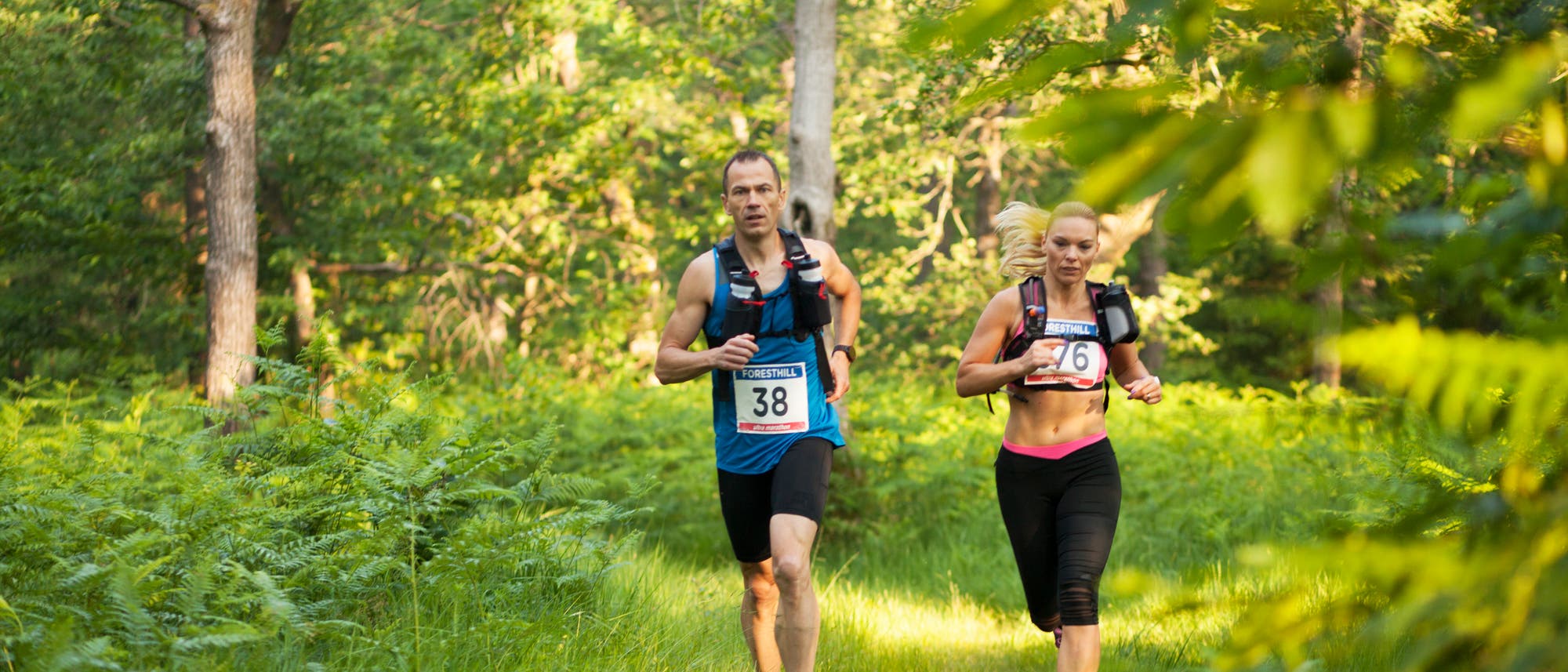 Eine Athletin und ein Athlet, die während eines Ultramarathons durch einen Waldweg laufen.