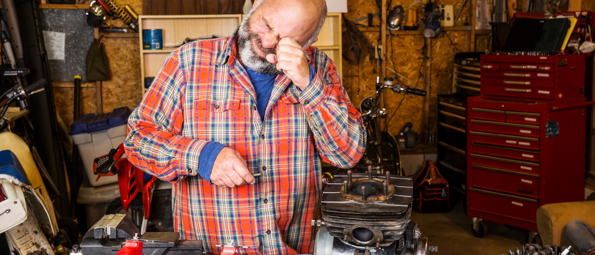 Ein Mann repariert etwas in seiner Werkstatt und hält sich sein linkes Auge mit der Hand zu, weil er vermutlich etwas ins Auge bekommen hat.