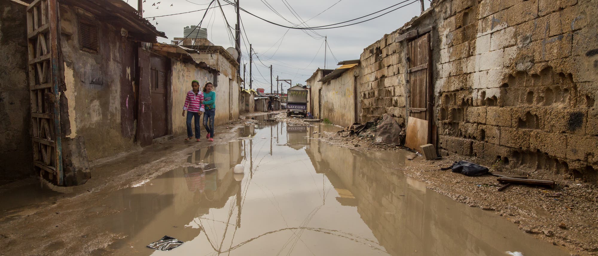 Wasser steht in einer Straße zwischen ärmlichen Behausungen.
