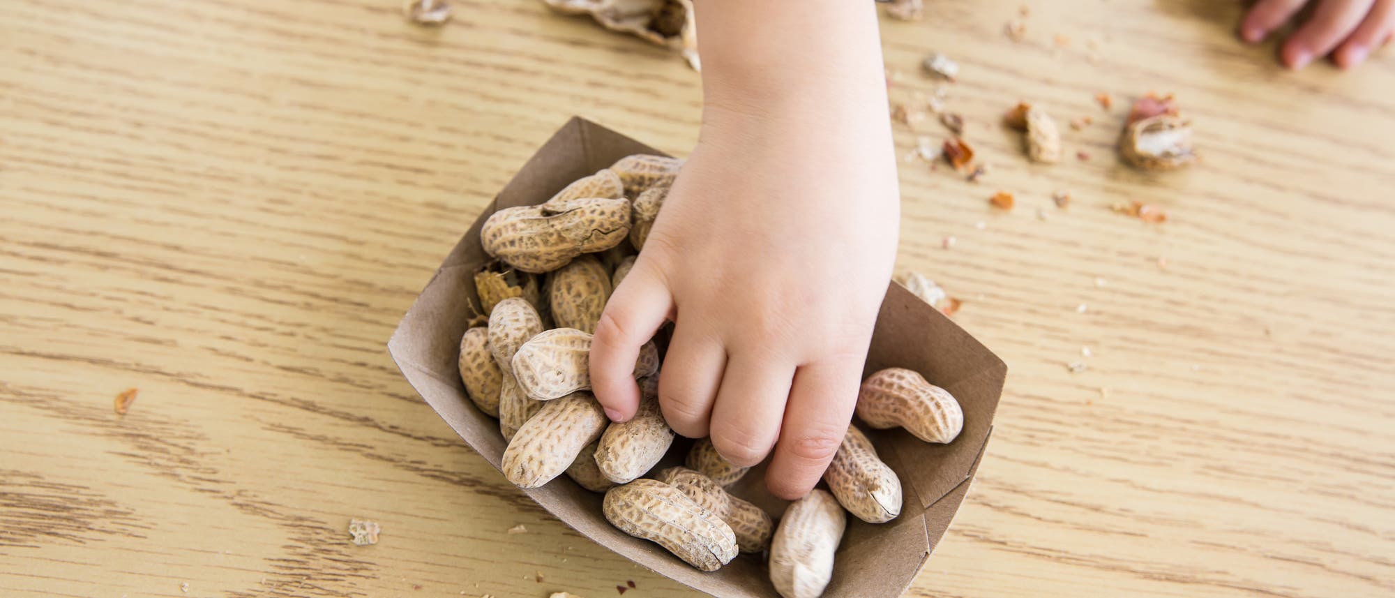Draufsicht auf eine Kinderhand, die in ein Schälchen voller Erdnüsse greift, das auf einem Tisch steht. 