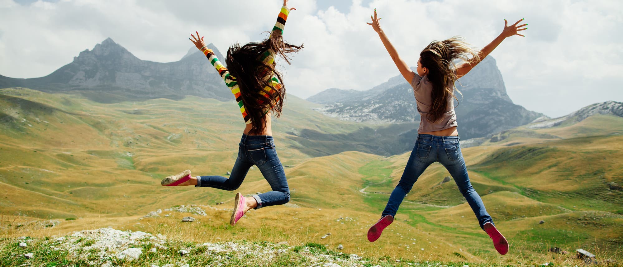 Zwei energiegeladene Mädchen springen vor einer Bergkulisse in die Höhe 