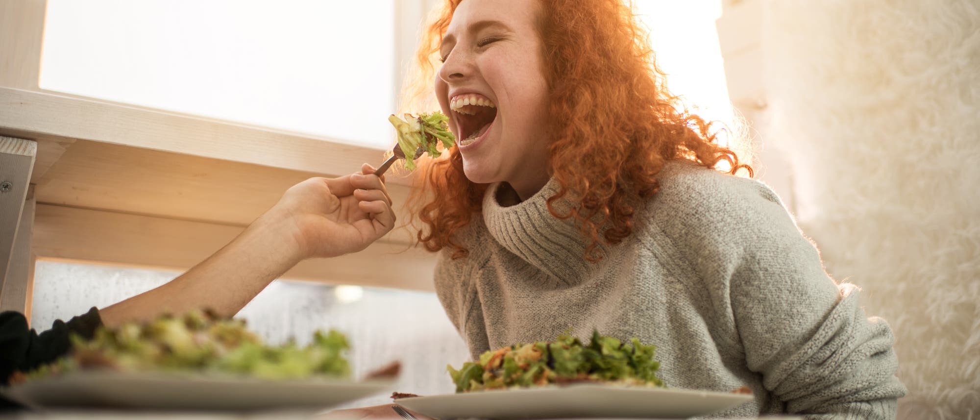 Eine jungen Frau mit roten Haaren wird von einem Mann mit Salat gefüttert. Sie lacht.