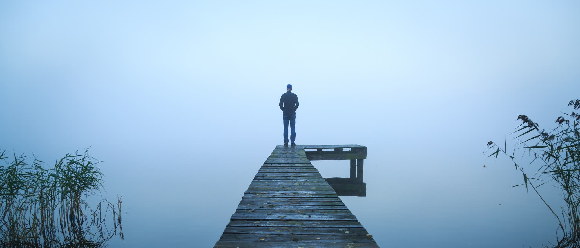 Mann steht am Ende eines Stegs und blickt auf eine neblige Seenlandschaft
