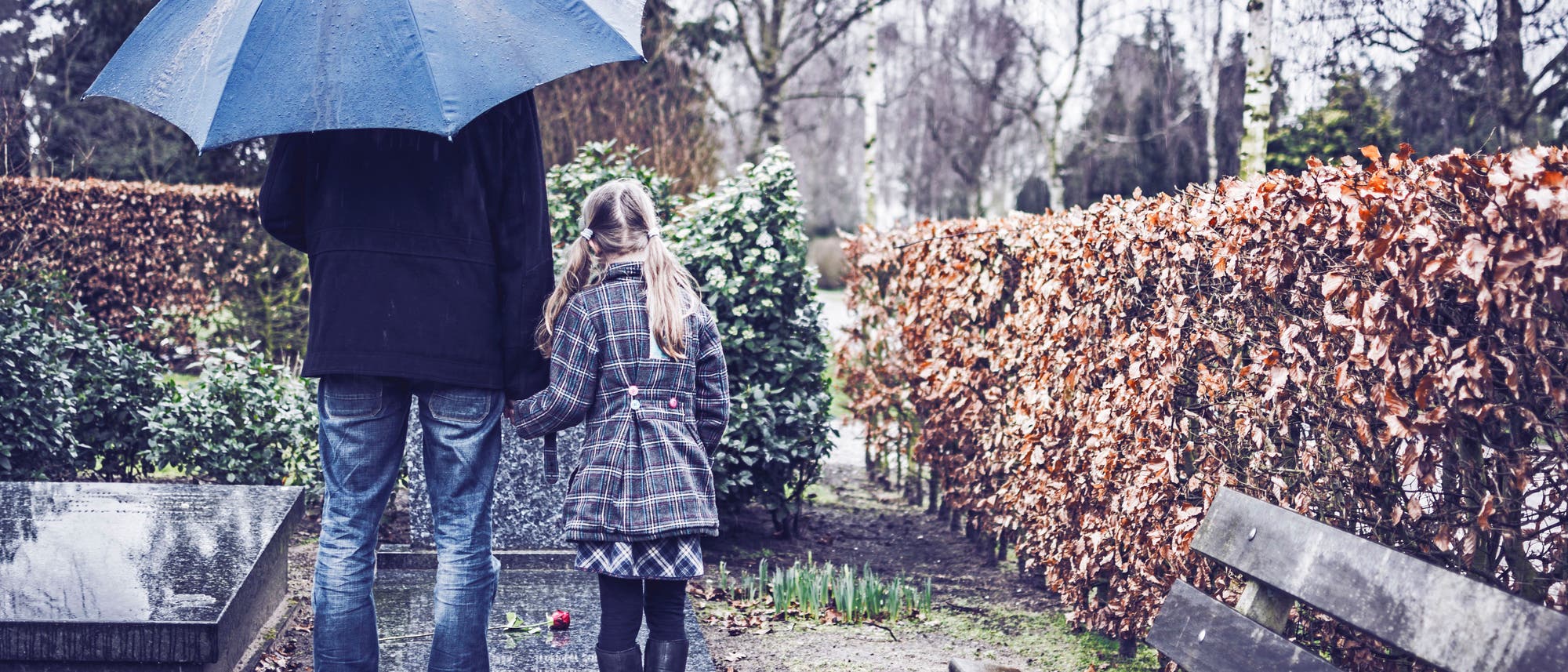 Ein Erwachsener und ein Kind stehen im Regen vor einem Grab mit Rose auf der Grabplatte..