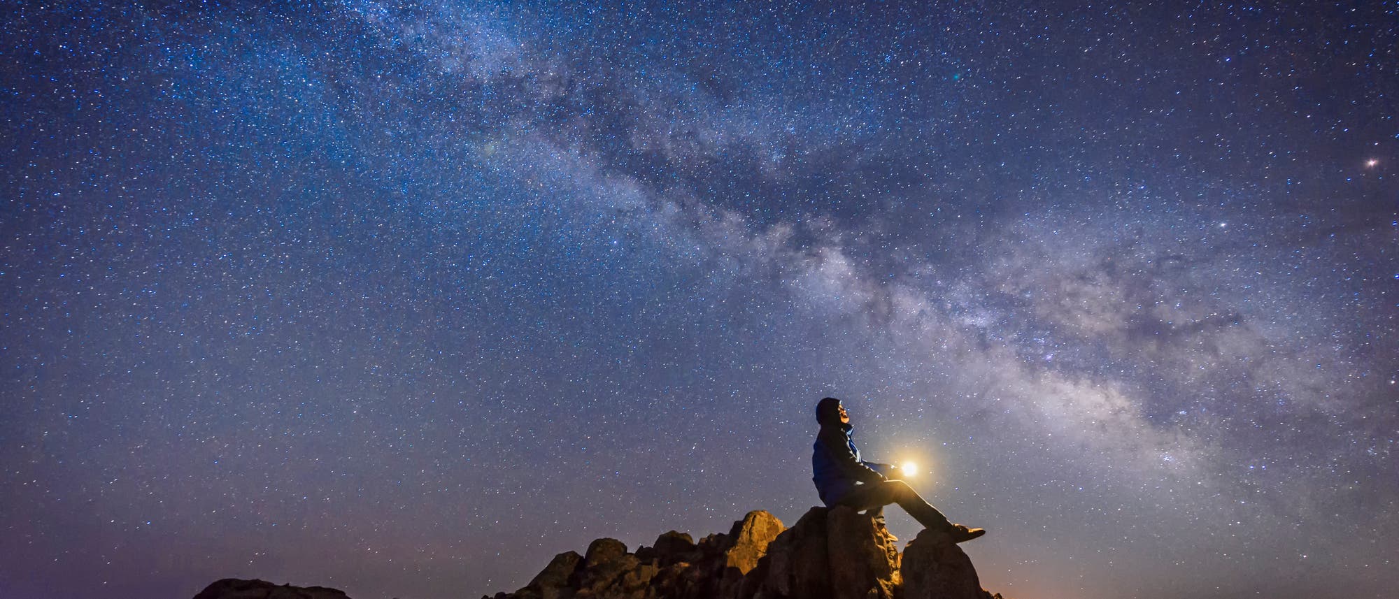 Ein Mann sitzt auf einem Fels und schaut auf die Milchstraße