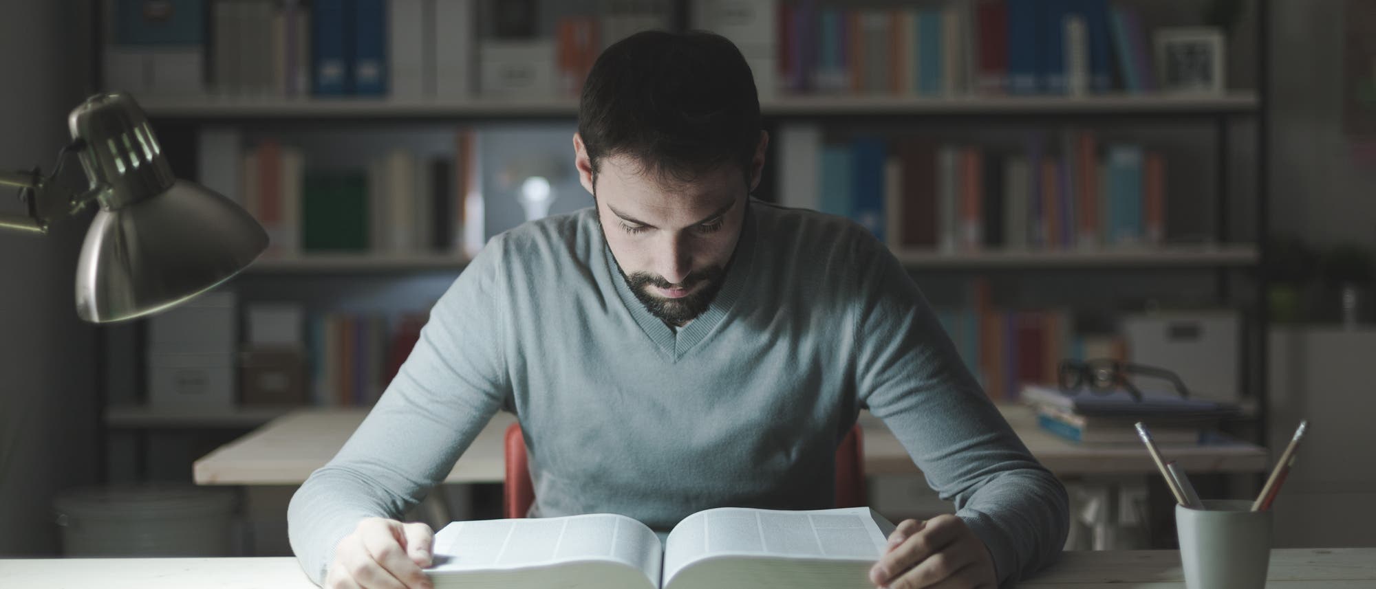 Ein Mann blickt konzentriert in ein dickes Buch. Hinter ihm steht ein Bücherregal. Eine Schreibtischlampe beleuchtet das Buch.
