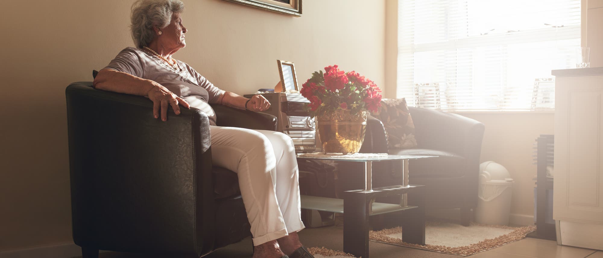 Eine Seniorin sitzt in einem Sessel, im Hintergrund ein hell erleuchtetes Fenster