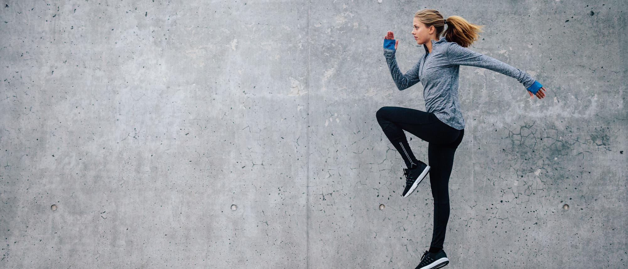 Eine Person in Sportkleidung springt dynamisch vor einer grauen Betonwand. Sie trägt eine graue Jacke, schwarze Leggings und schwarze Sportschuhe. Die Haare sind zu einem Pferdeschwanz gebunden. Die Szene vermittelt Energie und Bewegung.