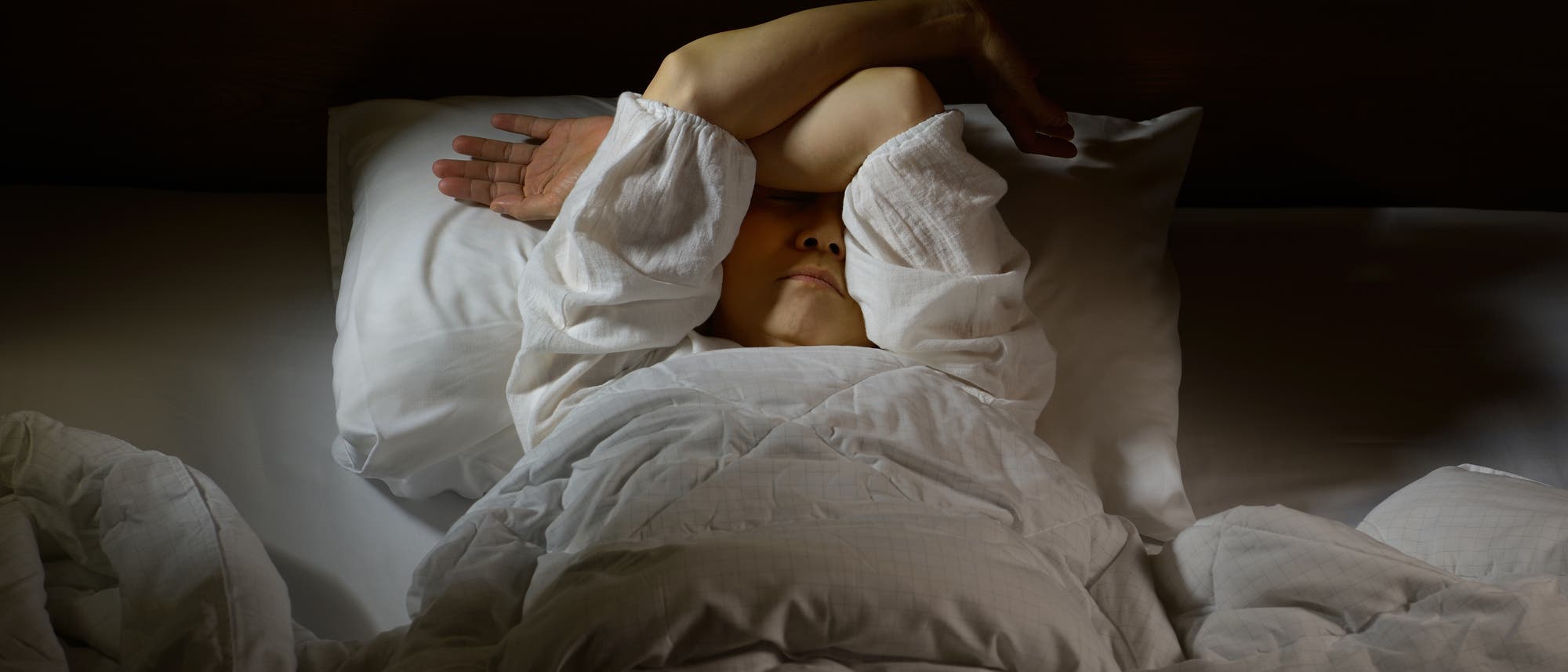 Eine Frau liegt unter einer Bettdecke und verschränkt die Arme vor ihrem Gesicht.