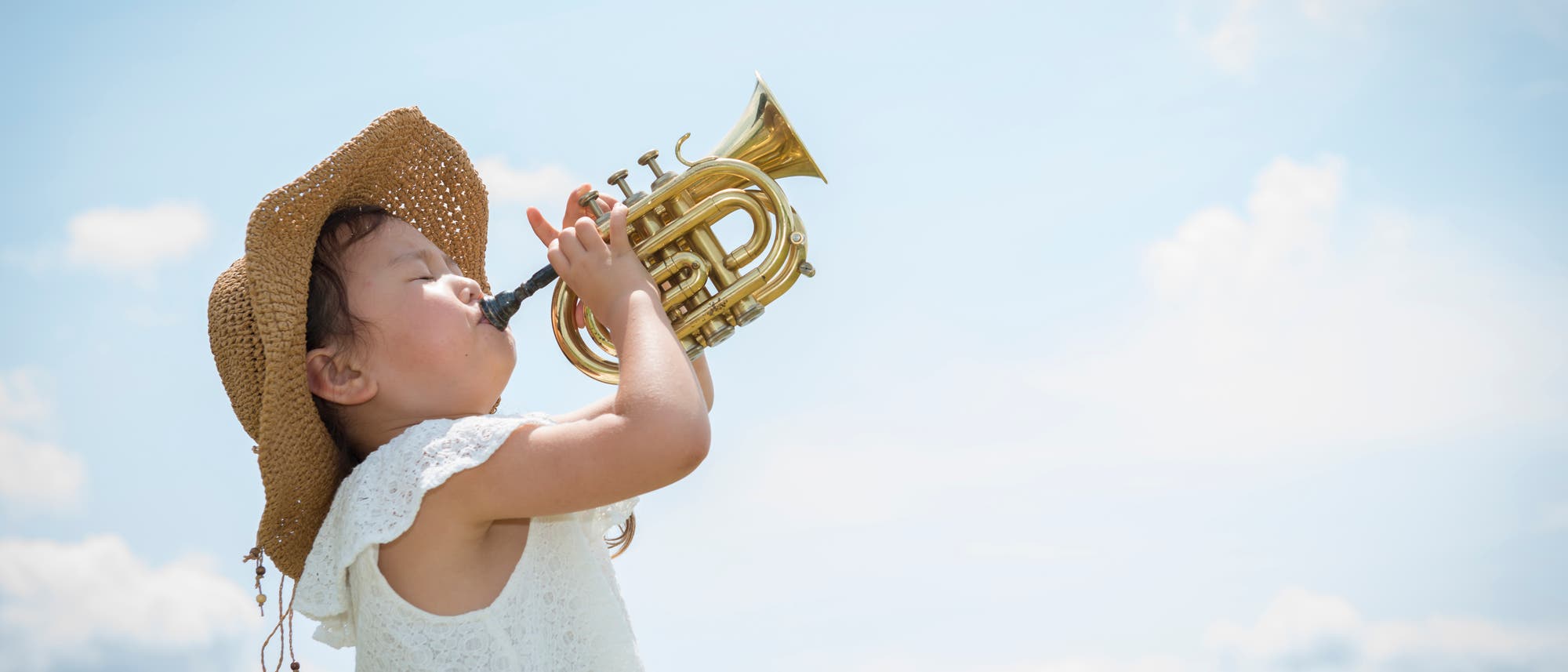 Ein kleines Mädchen mit weißem Kleid steht vor einem blauen Himmel und bläst in eine kleine Trompete.