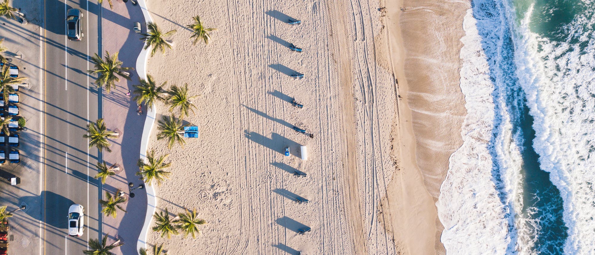 Das Bild zeigt einen Strand, an den rechts Wellen branden, während links eine Straße entlang läuft. Am Sandstrand selbst finden sich Rillen und Spuren im Sand.