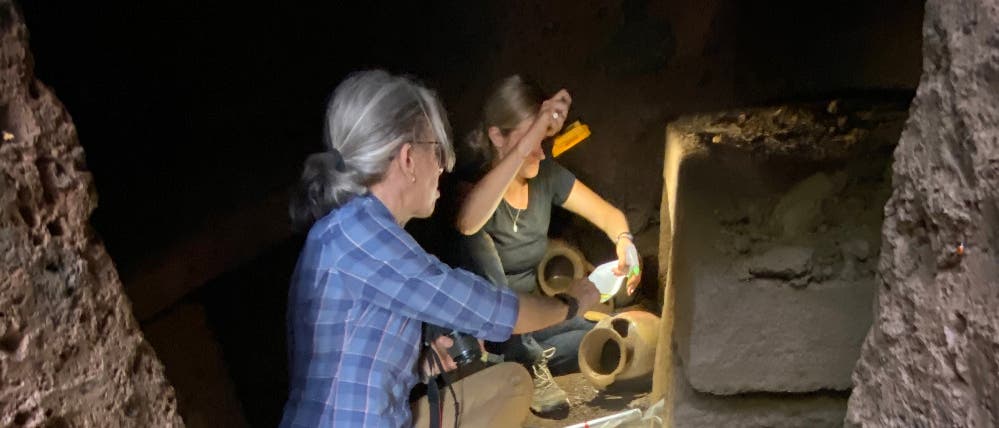 Zwei Personen arbeiten in einer archäologischen Ausgrabungsstätte. Sie befinden sich in einem dunklen Raum, der von einer Taschenlampe beleuchtet wird. Im Hintergrund sind antike Keramiken und Steine zu sehen. Eine Person hält ein Werkzeug, während die andere etwas untersucht. Die Szene vermittelt den Eindruck von konzentrierter Forschung und Entdeckung.