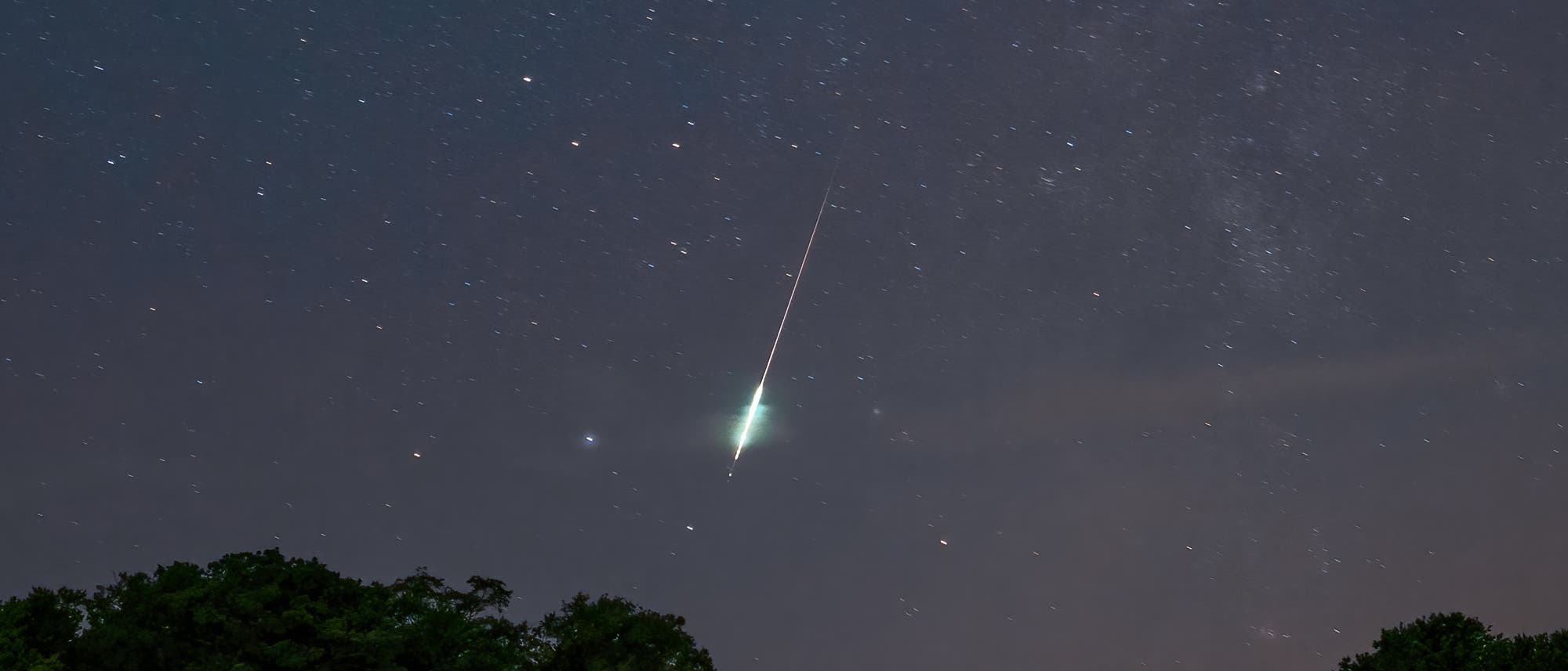 Ein Nachthimmel mit zahlreichen Sternen, durch den ein heller Meteor mit einem leuchtenden Schweif zieht. Im Vordergrund sind die Silhouetten von B&auml;umen und einer Wiese zu sehen. Der Meteor erhellt einen Teil des Himmels und zieht eine diagonale Linie von oben nach unten.