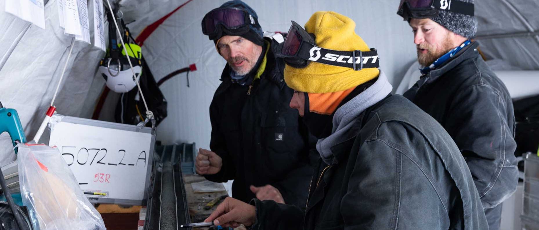 Drei Personen in Winterkleidung arbeiten in einem Zelt an einer wissenschaftlichen Untersuchung. Sie betrachten einen langen, zylindrischen Gegenstand auf einem Tisch, einen Bohrkern. Auf dem Tisch liegen Werkzeuge und ein Klemmbrett. An der Wand hängt ein Schild mit der Aufschrift "5072.2_A". Die Personen tragen Schutzbrillen und Mützen. Die Umgebung wirkt kalt und funktional. Drei Personen in Winterkleidung arbeiten in einem Zelt an einer wissenschaftlichen Untersuchung. Sie betrachten einen langen, zylindrischen Gegenstand auf einem Tisch, einen Bohrkern. Auf dem Tisch liegen Werkzeuge und ein Klemmbrett. An der Wand hängt ein Schild mit der Aufschrift "5072.2_A". Die Personen tragen Schutzbrillen und Mützen. Die Umgebung wirkt kalt und funktional.