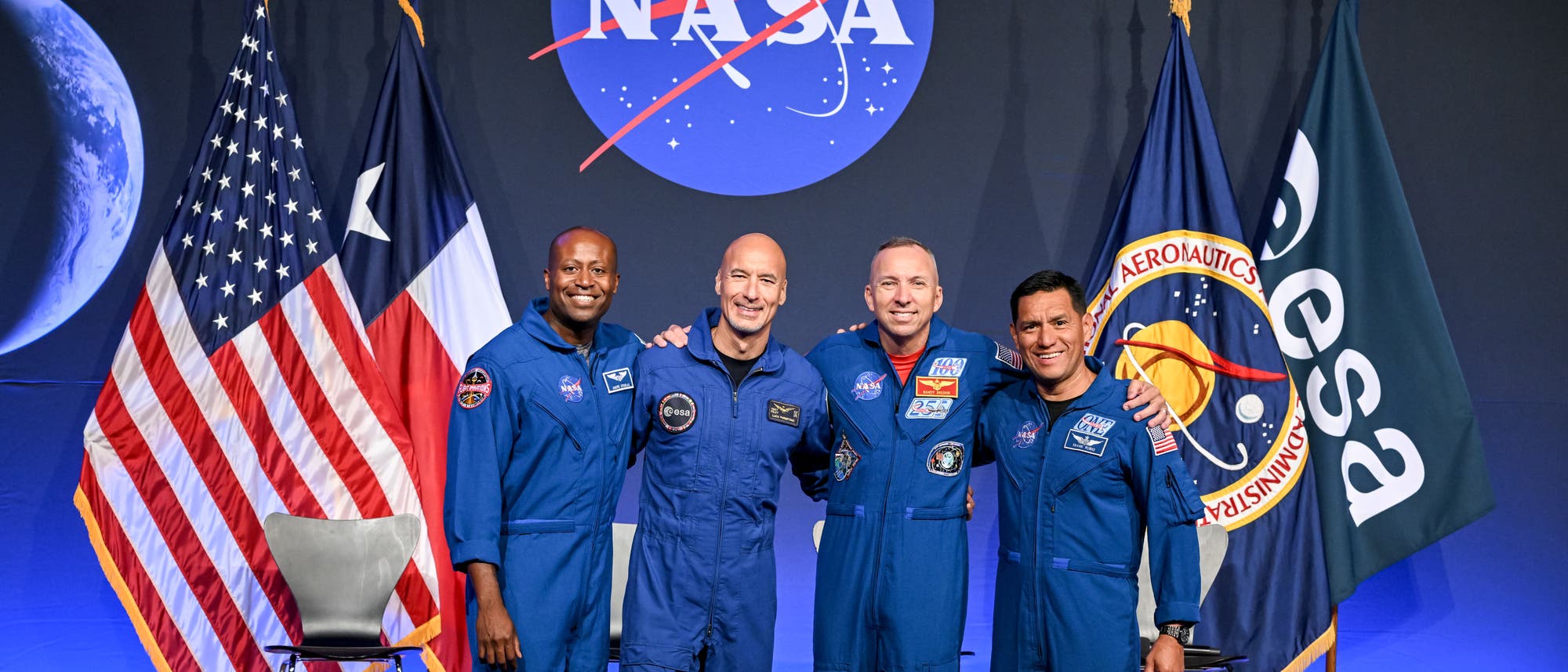 Vier Astronauten in blauen Raumanz&uuml;gen stehen l&auml;chelnd nebeneinander vor einem Hintergrund mit dem NASA-Logo. Links und rechts von ihnen sind die Flaggen der USA und der ESA zu sehen. Im Hintergrund ist ein Bild des Mondes zu erkennen. Die Astronauten haben ihre Arme umeinandergelegt und wirken fr&ouml;hlich und kameradschaftlich.