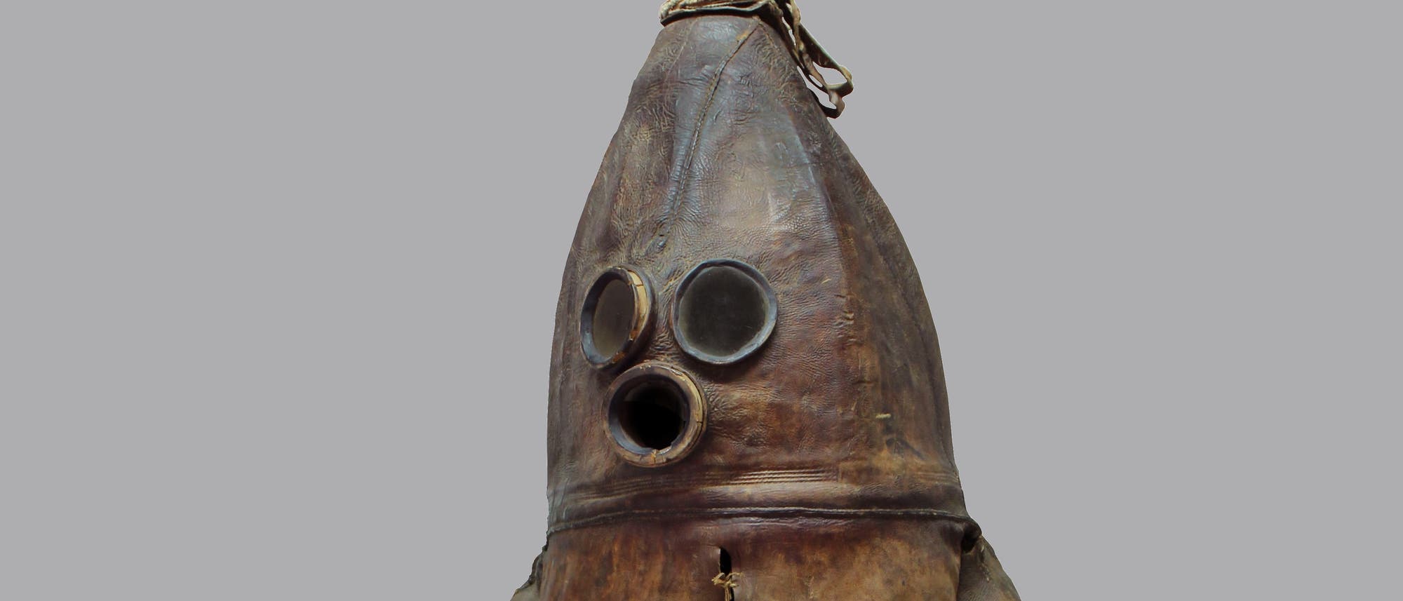 Eine alte Taucherausrüstung aus Leder, die einem Anzug ähnelt. Der Anzug hat eine konische Form mit zwei runden Glasfenstern für die Augen und einem runden Atemloch. Oben befindet sich eine Öffnung mit einem Verschluss, der mit einer Schnur gesichert ist. Der Hintergrund ist einfarbig grau. Eine alte Taucherausrüstung aus Leder, die einem Anzug ähnelt. Der Anzug hat eine konische Form mit zwei runden Glasfenstern für die Augen und einem runden Atemloch. Oben befindet sich eine Öffnung mit einem Verschluss, der mit einer Schnur gesichert ist. Der Hintergrund ist einfarbig grau.