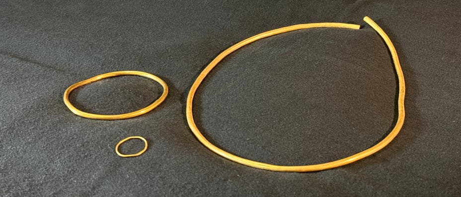 Drei goldene Schmuckstücke liegen auf einem dunklen Hintergrund. Links oben befindet sich ein mittelgroßer Armreif, darunter ein kleiner Ring. Rechts ist ein großer, offener Halsreif zu sehen. Die Schmuckstücke sind schlicht und glänzen im Licht. Drei goldene Schmuckstücke liegen auf einem dunklen Hintergrund. Links oben befindet sich ein mittelgroßer Armreif, darunter ein kleiner Ring. Rechts ist ein großer, offener Halsreif zu sehen. Die Schmuckstücke sind schlicht und glänzen im Licht.