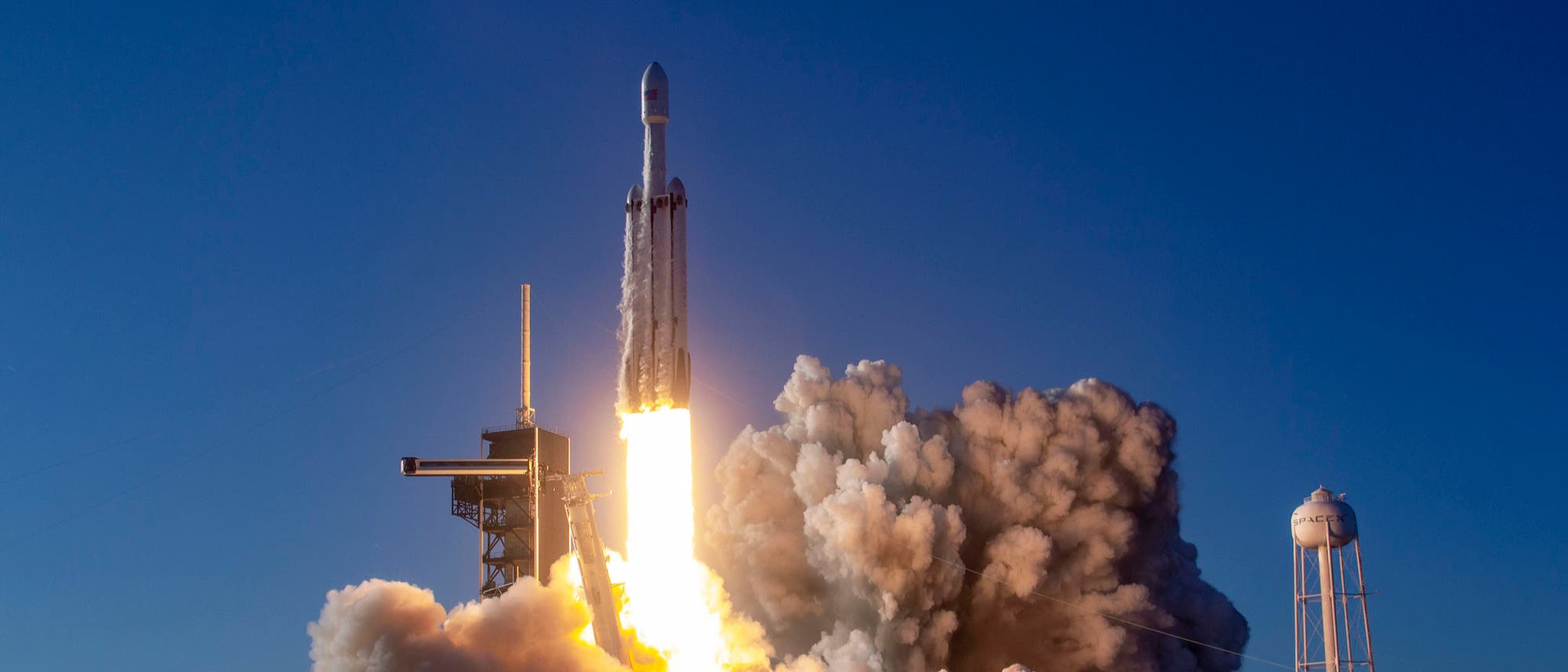 Eine Rakete hebt von einer Startrampe ab, umgeben von dichten Rauchwolken und hellem Feuerstrahl. Der Himmel ist klar und blau. Rechts im Bild ist ein hoher Wasserturm zu sehen. Die Szene vermittelt den kraftvollen Moment des Raketenstarts.