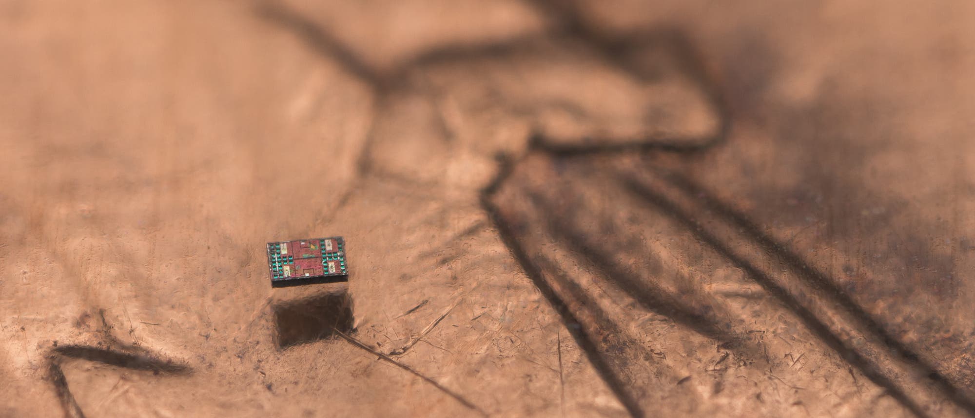 Ein winziger Mikrochip liegt auf einer strukturierten, metallischen Oberfl&auml;che. Der Chip zeigt komplexe Schaltkreise und bunte Komponenten. Die Oberfl&auml;che hat eine ungleichm&auml;&szlig;ige Textur mit Linien und Vertiefungen, die im Hintergrund verlaufen. Das Bild hebt die Miniaturgr&ouml;&szlig;e des Chips im Vergleich zur Oberfl&auml;che hervor.