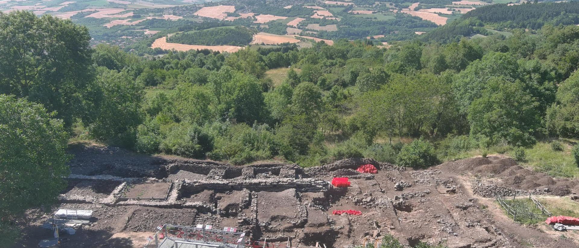 Luftaufnahme einer archäologischen Ausgrabungsstätte in einer hügeligen Landschaft. Im Vordergrund sind die Überreste von Steinmauern und rechteckigen Strukturen zu sehen, umgeben von Bäumen und Vegetation. Ein kleiner Bagger und rote Markierungen sind auf dem Gelände sichtbar. Im Hintergrund erstreckt sich eine weite Landschaft mit Feldern und Wäldern bis zu den entfernten Hügeln am Horizont. Luftaufnahme einer archäologischen Ausgrabungsstätte in einer hügeligen Landschaft. Im Vordergrund sind die Überreste von Steinmauern und rechteckigen Strukturen zu sehen, umgeben von Bäumen und Vegetation. Ein kleiner Bagger und rote Markierungen sind auf dem Gelände sichtbar. Im Hintergrund erstreckt sich eine weite Landschaft mit Feldern und Wäldern bis zu den entfernten Hügeln am Horizont.