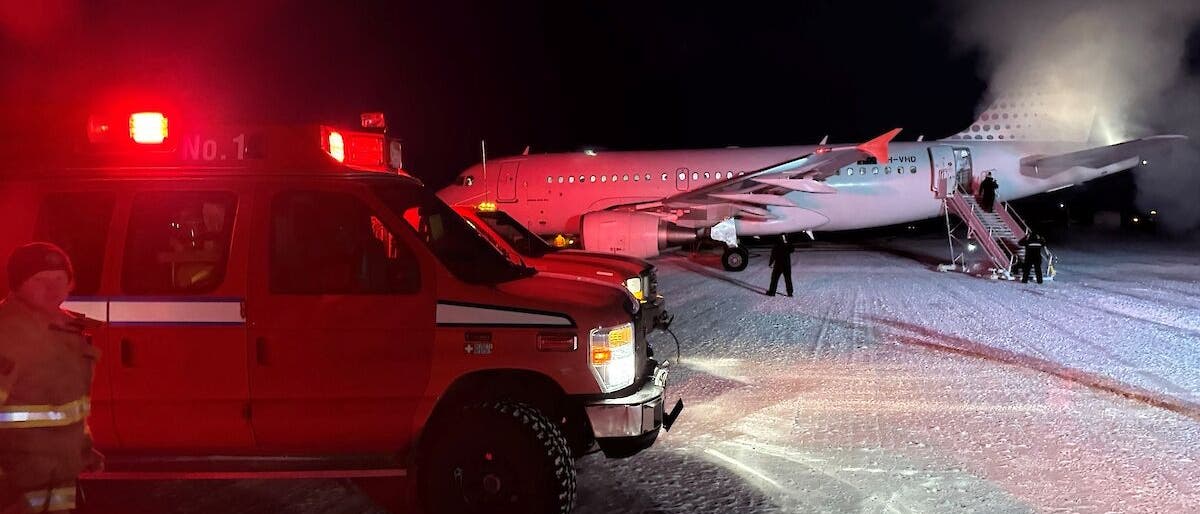 Ein Flugzeug steht nachts auf einer schneebedeckten Landebahn. Im Vordergrund ist ein rotes Einsatzfahrzeug mit eingeschaltetem Warnlicht zu sehen. Personen steigen über eine Treppe in das Flugzeug ein oder aus. Rauch oder Dampf steigt aus dem Flugzeug auf, was auf kalte Temperaturen hinweist. Die Szene wird von den Lichtern des Einsatzfahrzeugs beleuchtet. Ein Flugzeug steht nachts auf einer schneebedeckten Landebahn. Im Vordergrund ist ein rotes Einsatzfahrzeug mit eingeschaltetem Warnlicht zu sehen. Personen steigen über eine Treppe in das Flugzeug ein oder aus. Rauch oder Dampf steigt aus dem Flugzeug auf, was auf kalte Temperaturen hinweist. Die Szene wird von den Lichtern des Einsatzfahrzeugs beleuchtet.