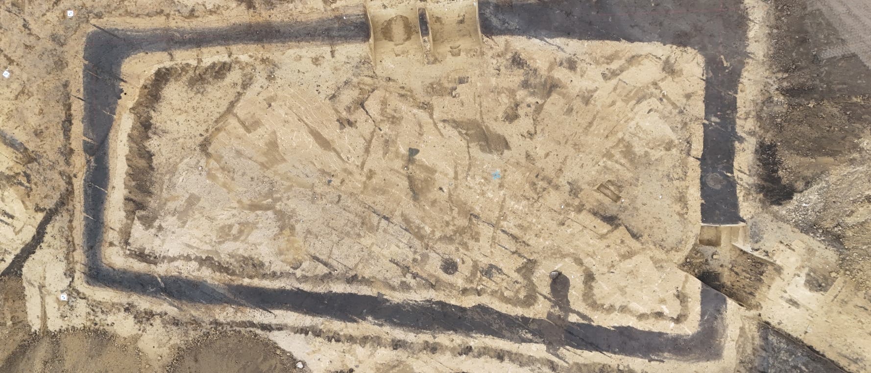 Luftaufnahme einer arch&auml;ologischen Ausgrabungsst&auml;tte mit rechteckigen Umrissen. Die Fl&auml;che ist von dunklen Linien umgeben, die Gr&auml;ben darstellen. Der Boden zeigt verschiedene Schattierungen und Texturen, die auf unterschiedliche Erdschichten oder Materialien hinweisen k&ouml;nnten. Die Umgebung ist von Erde und Vegetation umgeben.