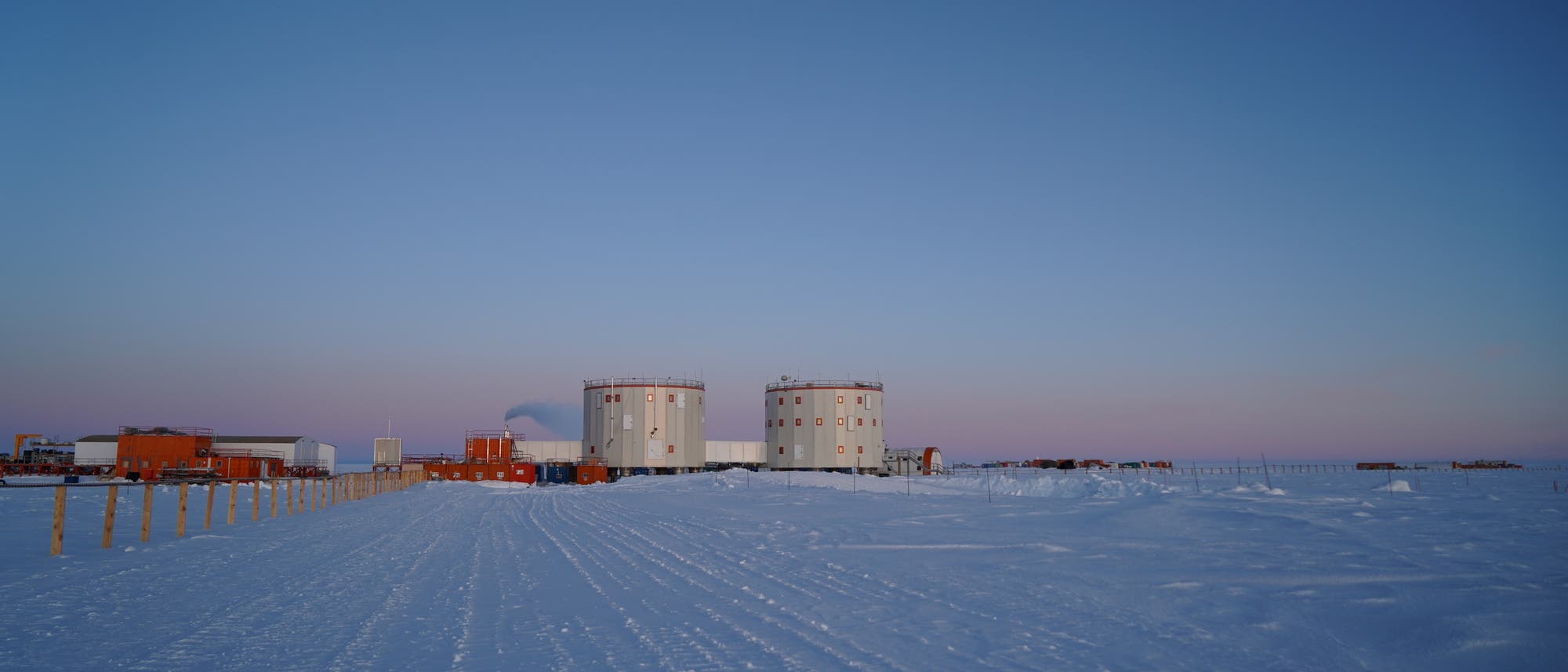 Eine Forschungsstation in einer verschneiten, arktischen Landschaft bei Sonnenuntergang. Zwei große, zylindrische Gebäude stehen im Zentrum, umgeben von kleineren Strukturen. Der Himmel ist klar mit einem sanften Farbverlauf von Blau zu Rosa. Schneebedeckte Wege führen zu den Gebäuden, und im Hintergrund sind weitere Einrichtungen sichtbar. Eine Forschungsstation in einer verschneiten, arktischen Landschaft bei Sonnenuntergang. Zwei große, zylindrische Gebäude stehen im Zentrum, umgeben von kleineren Strukturen. Der Himmel ist klar mit einem sanften Farbverlauf von Blau zu Rosa. Schneebedeckte Wege führen zu den Gebäuden, und im Hintergrund sind weitere Einrichtungen sichtbar.