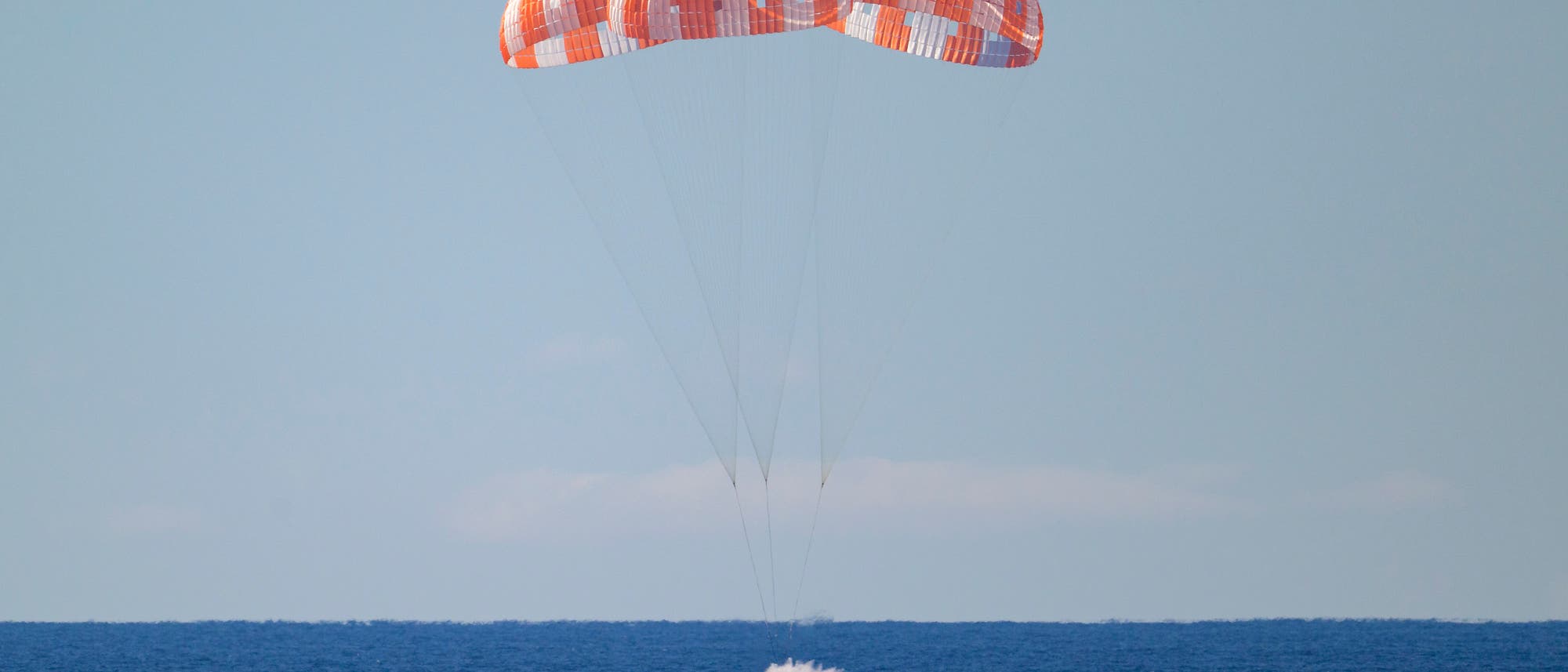 Die Raumkapsel Orion landet sicher im Ozean, gebremst von drei gro&szlig;en, rot-wei&szlig;en Fallschirmen. Die Kapsel erzeugt eine sichtbare Wasserfont&auml;ne beim Aufprall auf die Wasseroberfl&auml;che. Der Himmel ist klar und blau.