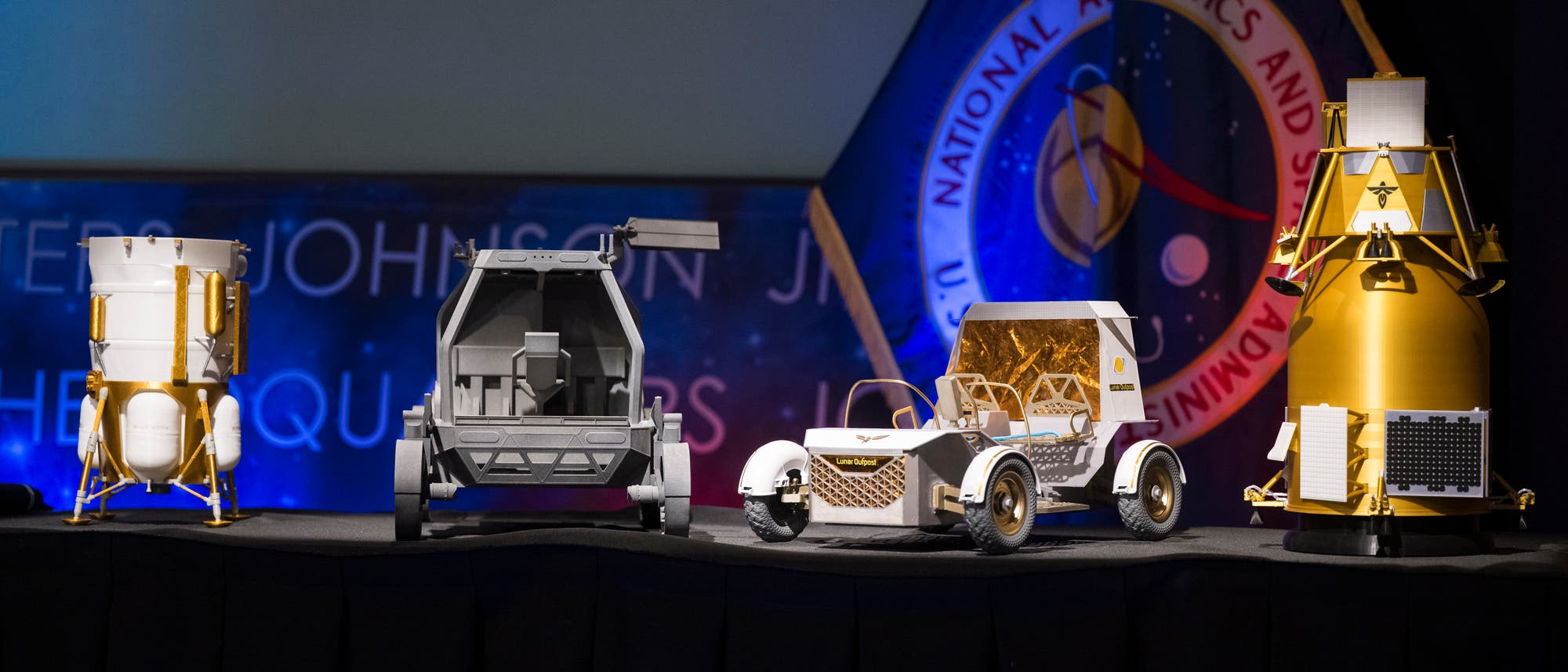 Eine Ausstellung von vier Modellen von Raumfahrzeugen auf einem Tisch. Die Modelle umfassen verschiedene Designs von Landefähren und Rovern, die in silbernen und goldenen Farben gehalten sind. Im Hintergrund ist ein Banner mit dem Logo einer Raumfahrtorganisation zu sehen. Die Szene ist in einem gut beleuchteten Raum mit einem dunklen Vorhang im Hintergrund. Eine Ausstellung von vier Modellen von Raumfahrzeugen auf einem Tisch. Die Modelle umfassen verschiedene Designs von Landefähren und Rovern, die in silbernen und goldenen Farben gehalten sind. Im Hintergrund ist ein Banner mit dem Logo einer Raumfahrtorganisation zu sehen. Die Szene ist in einem gut beleuchteten Raum mit einem dunklen Vorhang im Hintergrund.