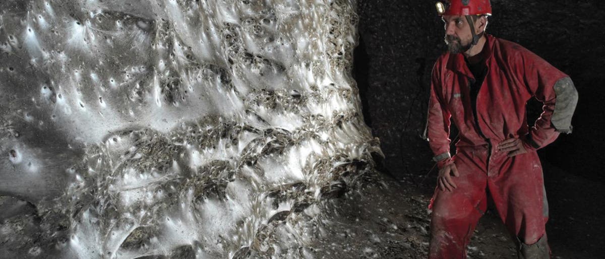 Ein H&ouml;hlenforscher in roter Schutzkleidung und Helm mit Stirnlampe steht in einer dunklen H&ouml;hle vor einer wei&szlig;-gl&auml;nzend versponnenen Felsformation. Die Oberfl&auml;che der Formation ist unregelm&auml;&szlig;ig und reflektiert das Licht, wodurch ein schimmernder Effekt entsteht. Der Forscher betrachtet die Struktur aufmerksam.