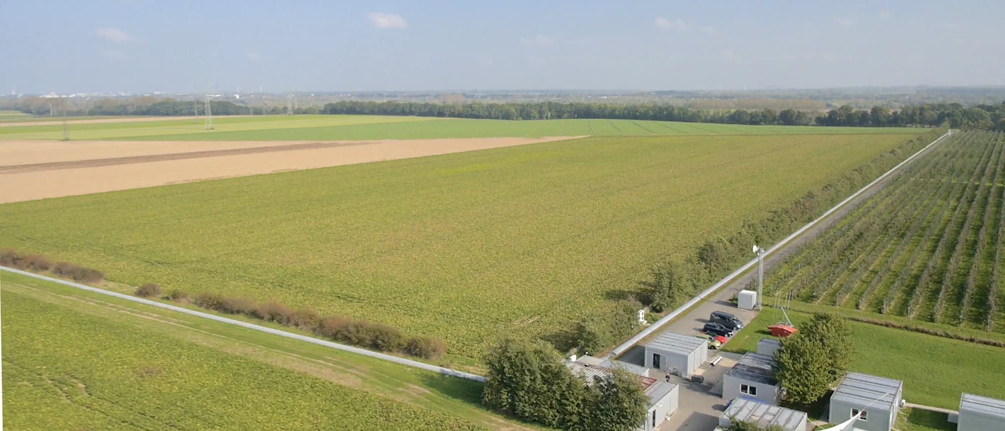 Luftaufnahme einer weitläufigen landwirtschaftlichen Landschaft mit Feldern in verschiedenen Grüntönen. Im Vordergrund befindet sich eine kleine Ansammlung von Gebäuden und Fahrzeugen, die von Bäumen umgeben sind. Ein schmaler Weg verläuft entlang der Felder. Der Himmel ist klar mit wenigen Wolken. Luftaufnahme einer weitläufigen landwirtschaftlichen Landschaft mit Feldern in verschiedenen Grüntönen. Im Vordergrund befindet sich eine kleine Ansammlung von Gebäuden und Fahrzeugen, die von Bäumen umgeben sind. Ein schmaler Weg verläuft entlang der Felder. Der Himmel ist klar mit wenigen Wolken.