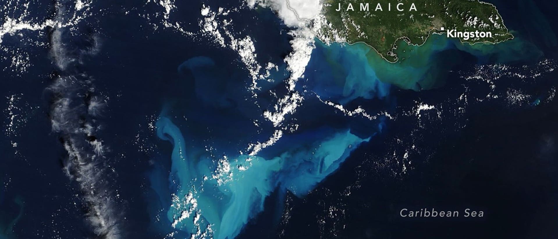 Satellitenbild der Karibik, das die Insel Jamaika zeigt. Die Karte hebt die K&uuml;stenlinie und die Stadt Kingston hervor. Umgeben von tiefblauem Wasser, sind helle Wolkenformationen und t&uuml;rkisfarbene Str&ouml;mungen im Meer sichtbar. Der Text "Jamaica" und "Caribbean Sea" ist auf der Karte vermerkt.