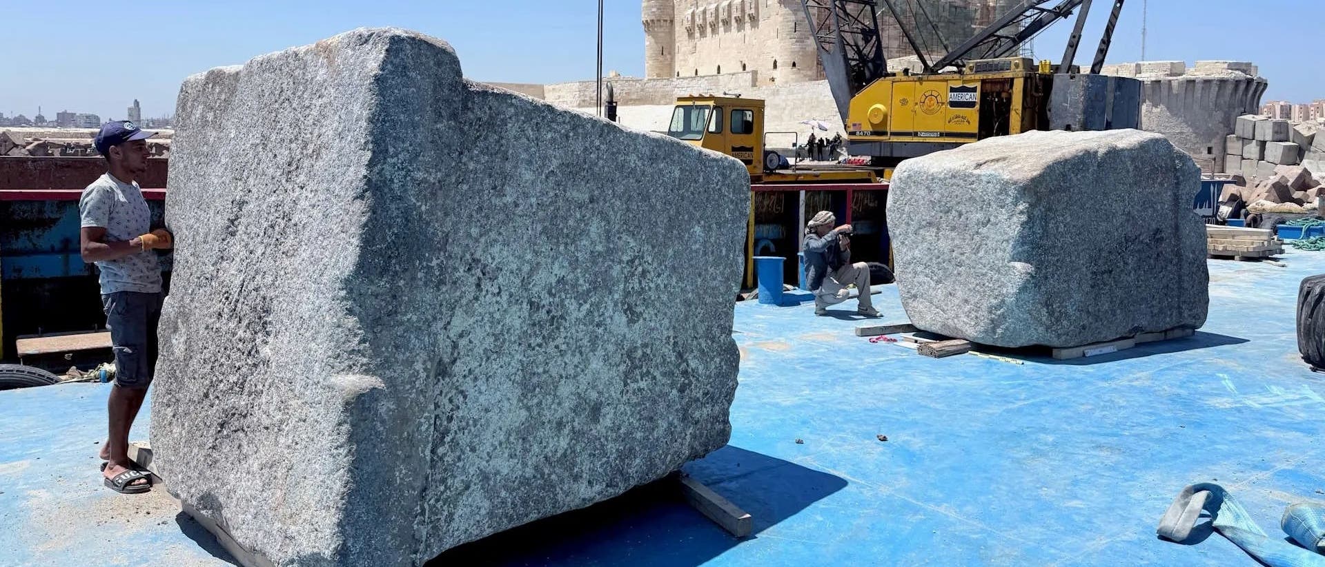 Ein Arbeiter steht neben einem großen, rechteckigen Steinblock an einer Stelle mit blauem Bodenbelag. Im Hintergrund ist ein gelber Kran zu sehen, der einen weiteren Steinblock hebt. Eine historische Festung erhebt sich im Hintergrund unter einem klaren blauen Himmel. Eine ägyptische Flagge weht auf dem Kran. Die Szene vermittelt den Eindruck von archäologischen Arbeiten in der Nähe eines historischen Ortes. Ein Arbeiter steht neben einem großen, rechteckigen Steinblock an einer Stelle mit blauem Bodenbelag. Im Hintergrund ist ein gelber Kran zu sehen, der einen weiteren Steinblock hebt. Eine historische Festung erhebt sich im Hintergrund unter einem klaren blauen Himmel. Eine ägyptische Flagge weht auf dem Kran. Die Szene vermittelt den Eindruck von archäologischen Arbeiten in der Nähe eines historischen Ortes.